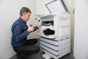 man kneeling and holding screw driver while inspecting copier machine