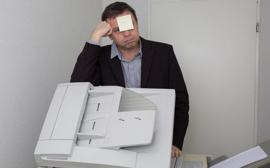 A man sitting at a business copier machine with a post it note on his forehead saying out of order