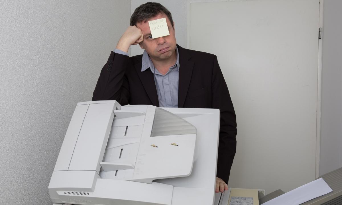 A man sitting at a business copier machine with a post it note on his forehead saying out of order