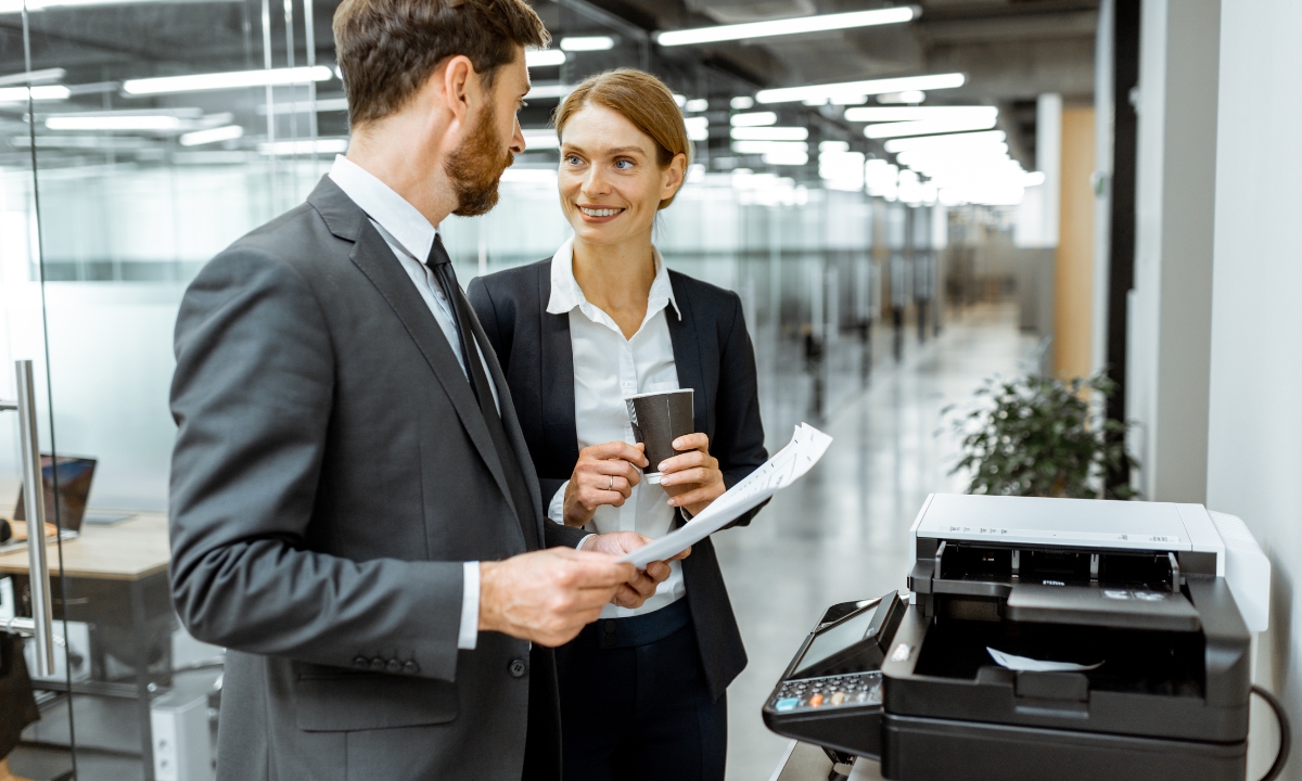 two coworkers talking and drinking coffee by a photocopier