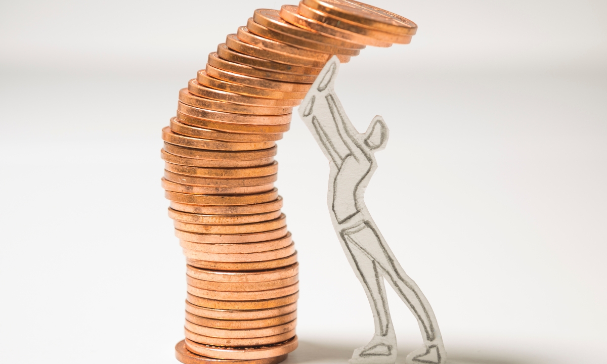 a small paper person holding up a falling stack of metal coins